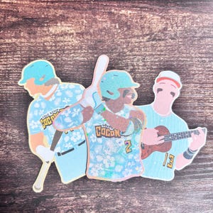 Puede incluir: Tres pegatinas con temática de béisbol. Una pegatina muestra a un jugador de béisbol con un bate, con un casco y una camiseta azules con el texto "Loco Beach Cocon" y el número 2. Otra pegatina muestra a un jugador de béisbol con bigote. La tercera muestra a un jugador de béisbol con un ukelele.
