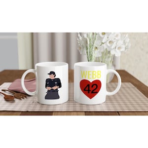 May include: Two white ceramic mugs featuring graphic designs. One mug displays a stylized illustration of a person in a dark uniform and cap. The other mug features the yellow text WEBB above a large red heart containing the black number 42.