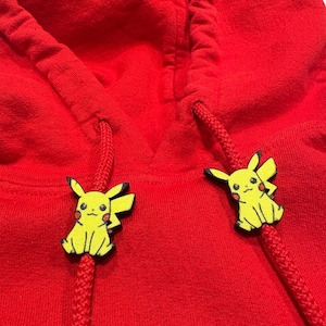 May include: Close-up of a red hoodie with yellow Pikachu-shaped cord ends. The Pikachu figures have black outlines and details. The hoodie's drawstring is red.