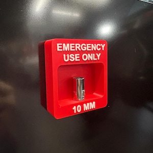 May include: A red plastic holder with the words "EMERGENCY USE ONLY" and "10 MM" printed in white. A silver metal socket is inside the holder. The holder is mounted on a black surface.