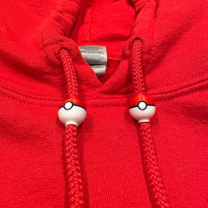 May include: Close-up of a red hooded sweatshirt with white and red ball-shaped drawstring ends. The drawstring is a thick, braided red cord. The sweatshirt's hood is visible in the background.