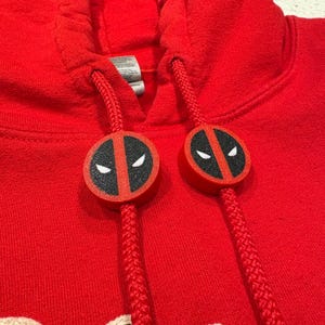 May include: A red hooded sweatshirt with a drawstring and Deadpool-themed accents. The drawstring ends feature circular red and black Deadpool face designs. The sweatshirt is made of a soft, textured material.