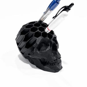 May include: A matte black, geometric skull-shaped desk organizer featuring a honeycomb-patterned top. The skull has a faceted, low-poly design and contains two pens, one with a blue cap and one with a white barrel, inserted into the hexagonal openings.