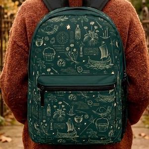 May include: A dark green backpack featuring a gold pattern of nautical and botanical illustrations. The backpack has black straps and a front pocket with a black zip. The person in the image is wearing a brown jumper.