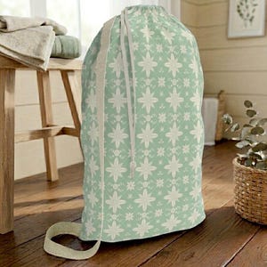 Puede incluir: Una bolsa de lavandería verde menta con un patrón floral blanco repetido. La bolsa tiene un cierre de cordón y una correa beige. La bolsa está sobre un suelo de madera. La altura de la bolsa es de unos 75 cm.