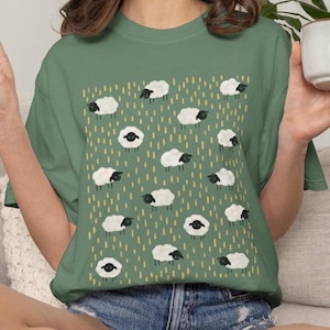 May include: A sage green short-sleeved t-shirt featuring a repeating pattern of small white sheep with black faces and legs, interspersed with vertical yellow dashes across the front of the garment, paired with blue denim shorts.