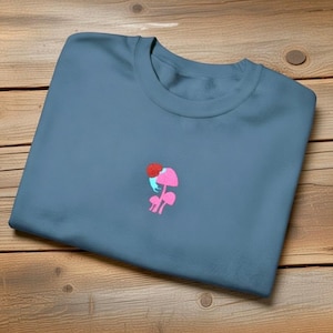 May include: A blue-grey crew neck sweatshirt. It has a small embroidered design of a pink mushroom with a red top and blue accents. The sweatshirt is folded on a wooden surface.
