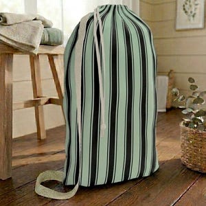 Puede incluir: Una bolsa de lavandería alta con cierre de cordón. La bolsa presenta rayas verticales en negro y verde claro. Una correa de color crema está unida al lateral de la bolsa. La bolsa está sobre un suelo de madera.