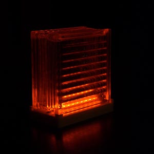 Può includere: Una luce rettangolare arancione incandescente con linee orizzontali, su sfondo nero. La luce sembra essere un piccolo dispositivo illuminato, forse una spia o un indicatore. Emette un bagliore ambrato caldo.