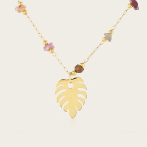 Può includere: Una collana color oro con un ciondolo centrale a forma di foglia di monstera sospeso a una catenina sottile. La catena è impreziosita da piccole perline dorate lucide e frammenti di pietra irregolari e multicolori nei toni del rosa, marrone e grigio.
