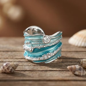 Puede incluir: Un anillo plateado con un diseño en forma de ola, con esmalte azul claro y piedras claras brillantes. Una sola piedra clara más grande está engastada en el centro. El anillo se exhibe sobre una superficie de madera con conchas marinas.