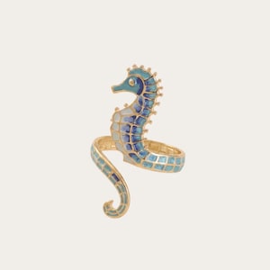 Könnte beinhalten: Ein goldfarbener Ring mit einem Seepferdchen-Motiv und blau-weißen Emaille-Details. Der Körper des Seepferdchens bildet den Mittelpunkt, wobei sich der Schwanz zu einem Ringband krümmt und ein segmentiertes, gitterartiges Muster aufweist.