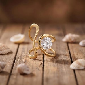 Può includere: Un anello color oro caratterizzato da un design aperto e sinuoso che forma un'asola asimmetrica. Una singola pietra rotonda e trasparente sfaccettata è incastonata al centro della parte circolare della fascia, creando un punto focale.