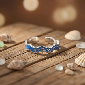 Può includere: Un anello aperto in argento caratterizzato da un design a fascia ondulata con un inserto in smalto blu. La superficie è decorata con piccoli motivi celesti color oro, tra cui una mezzaluna, stelle e linee vorticose lungo la fascia.