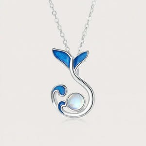 Può includere: Una collana in argento con un ciondolo a forma di coda di balena, decorato con smalto blu e una pietra di luna rotonda e iridescente. Il ciondolo è appeso a una sottile catenina d'argento e presenta una finitura metallica liscia e lucida.