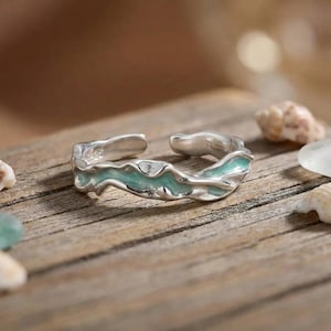May include: An open-ended silver ring featuring an irregular, wavy band design with a smooth, light seafoam green enamel inlay running along the centre of the metal surface, creating a fluid, organic appearance on the exterior of the band.