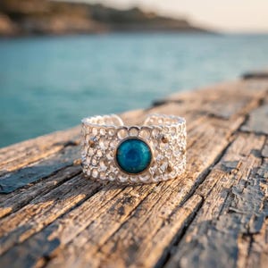 Può includere: Un anello color argento caratterizzato da una fascia larga e traforata con un motivo a nido d'ape composto da fori circolari. Una singola pietra blu, liscia e circolare, è incastonata al centro della fascia, circondata dalla struttura metallica.