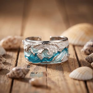 Può includere: Un anello aperto color argento con un design smaltato raffigurante onde oceaniche. L'anello presenta vortici di colori blu e bianco con piccole pietre trasparenti incastonate che imitano la schiuma marina e il movimento dell'acqua.