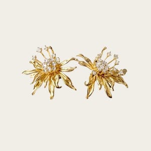 Può includere: Un paio di orecchini color oro a forma di fiori in piena fioritura con petali ondulati e testurizzati. Il centro di ogni fiore è raggruppato con piccole pietre chiare e scintillanti, con pietre aggiuntive sulle punte di sottili stami.
