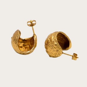 Peut inclure: Une paire de boucles d'oreilles créoles dorées avec une surface texturée et irrégulière. Chaque boucle présente un design épais en demi-cercle avec une finition polie et un mécanisme de fermeture à tige standard pour une fixation à l'oreille.
