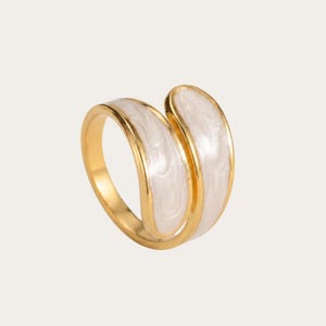 Può includere: Un anello color oro con un design aperto a bypass. La fascia è impreziosita da due inserti in smalto bianco madreperlaceo e liscio che seguono la forma curva del metallo, creando un aspetto fluido e continuo quando indossato al dito.