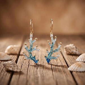 Può includere: Un paio di orecchini pendenti color oro a forma di corallo ramificato, con finitura in smalto blu, piccoli accenti bianchi e minuscole pietre trasparenti incastonate lungo i rami, sospesi a semplici ganci per orecchie dorati.