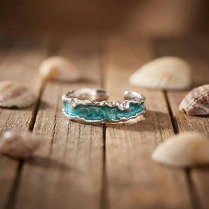 May include: A silver-toned open ring featuring a textured, wavy band with a vibrant turquoise-coloured inlay. The metal edges are irregular and organic in shape, creating a rugged appearance that contrasts with the smooth, bright blue-green centre.