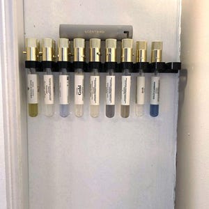 May include: A wall-mounted perfume sample holder with ten glass vials. Each vial has a gold-coloured cap and contains a different coloured liquid. The holder has the word "SCENTBIRD" printed on it.