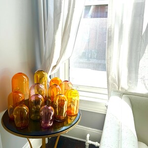 Könnte beinhalten: Eine Sammlung von Glasvasen in Orange-, Gelb- und Lilatönen steht auf einem runden Tisch. Der Tisch hat eine schwarze Platte und goldene Beine. Die Vasen haben verschiedene Formen und Größen. Sonnenlicht fällt durch ein Fenster.
