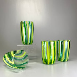 Puede incluir: Cuatro vasos de vidrio transparente para beber con diseños de rayas verdes y amarillas. Los vasos están dispuestos en una fila sobre una superficie blanca.