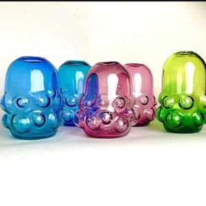 Puede incluir: Cinco jarrones de vidrio en diferentes colores, cada uno con un diseño texturizado único que se asemeja a un pulpo. Los jarrones están dispuestos en una fila sobre una superficie blanca.