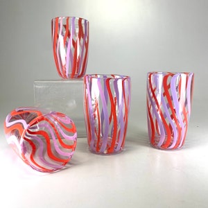 Puede incluir: Cuatro vasos de vidrio transparente con diseños de rayas rosas y naranjas. Los vasos están sobre una superficie blanca.