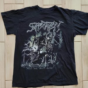 May include: Black t-shirt with the band name "Suffocation" in a gothic font above a skeletal graphic. The text "...Evil lies, death denied" is printed below the graphic. The shirt is made of a dark fabric.