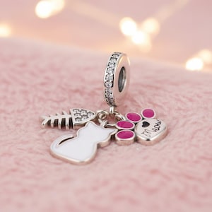 May include: A silver charm with a cat, fishbone, and paw print design. The charm features a white cat, a fishbone with clear accents, and a paw print with pink pads and the word "meow". The charm has a silver ring with clear stones.