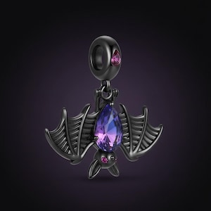 May include: A black bat charm featuring a purple teardrop-shaped gem. The bat's wings are outstretched, and it has small pink gems for eyes. A small pink gem is also set at the top of the charm.