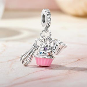 May include: A silver charm with a baking theme. It features a whisk, a cupcake with colourful gems, and a measuring cup. The charm has a loop at the top, encrusted with clear stones.