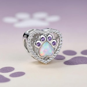 May include: A silver charm with a heart-shaped frame, encrusted with small clear stones. Inside, a paw print design features a white opal centre and purple accents. The charm is designed to be a pendant or bead.