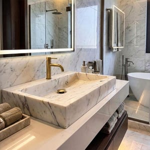 Puede incluir: Un baño moderno con un tocador de mármol y un lavabo rectangular. El grifo y el desagüe son dorados. El baño también cuenta con un espejo grande con luz, una ducha y una bañera. La combinación de colores es neutra, con tonos de blanco, gris y dorado.