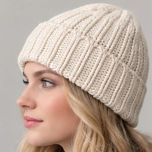 May include: A cream-colored knit beanie featuring a thick, vertical ribbed texture. The hat is constructed with a folded cuff at the base and a rounded crown, showcasing a consistent, chunky yarn pattern throughout the entire garment.