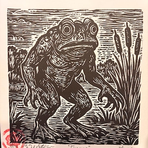 May include: Black and white linocut print of a large frog-like creature, titled "Frygmen". The artwork depicts the creature standing in a marshy environment with cattails and water in the background. Signed and numbered.