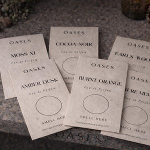 Puede incluir: Muestras de perfume OASES con nombres como "Cocoa Noir" y "Amber Dusk". Las tarjetas beige tienen texto negro y un área "Smell Here". El fondo es una superficie gris texturizada, con una vela encendida.