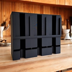 May include: A black, multi-compartment organizer, likely for kitchen or office use, stands on a wooden surface. The organizer has several vertical slots and lower compartments, suggesting storage for items like cups or utensils. The design is modern and functional.