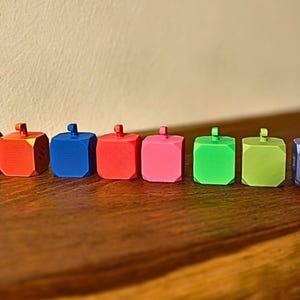May include: A row of twelve colorful, cube-shaped objects with small, cylindrical handles on top. The objects are in various colors, including black, white, blue, red, pink, green, purple, orange, and teal. They are arranged on a dark wooden surface.