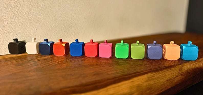 May include: A row of twelve colorful, cube-shaped objects with small, cylindrical handles on top. The objects are in various colors, including black, white, blue, red, pink, green, purple, orange, and teal. They are arranged on a dark wooden surface.