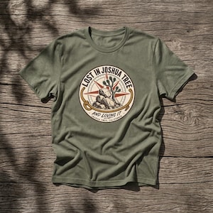 May include: An olive green short-sleeve t-shirt featuring a circular graphic on the chest. The design includes a compass rose, a Joshua tree, and rocks, surrounded by a rope border with the text LOST IN JOSHUA TREE AND LOVING IT JOSHUA TREE, CALIFORNIA.