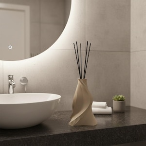 Könnte beinhalten: Ein beiger, skulpturaler Schilf-Diffusor mit schwarzen Schilfrohren, auf einer dunklen Arbeitsplatte in einem modernen Badezimmer. Ein weißes Waschbecken, ein Spiegel und eine kleine Topfpflanze sind ebenfalls zu sehen. Das Design des Diffusors hat eine Spiralform.