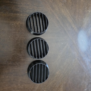 May include: Three black, circular air vent covers with vertical slats are arranged on a dark wooden surface. The vents have a rounded, recessed design, and the slats are evenly spaced.