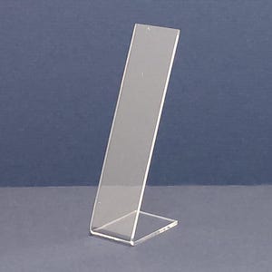 May include: A clear acrylic display stand with a slanted design. The stand has a rectangular base and a tall, angled panel for showcasing items. The stand is made of transparent material and is suitable for displaying various products.