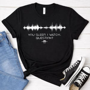 May include: Black t-shirt with white text and a sound wave graphic. The text reads "YOU SLEEP, I WATCH. QUESTION?" with a small alien-like symbol below. The shirt is paired with blue jeans and black sneakers.