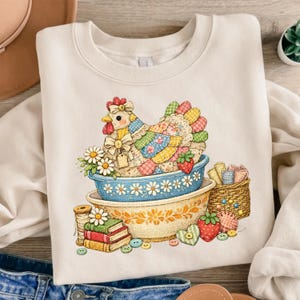 May include: Cream-colored sweatshirt featuring a colorful patchwork chicken design. The chicken sits in a blue and yellow bowl with daisies, strawberries, and other decorative elements. A stack of books and buttons are also visible.